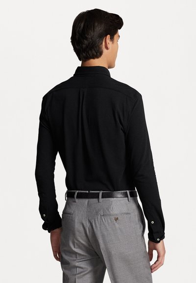 Zwarte langemouwen shirt met een klassieke kraag, van achteren bekeken. Gecombineerd met grijze op maat gemaakte broek en een zwarte riem. Gladde stofstructuur.
