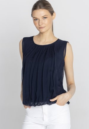 Kobieta o jasnej karnacji i blond włosach związanych nisko w kucyk, ubrana w granatową bezrękawnikową plisowaną bluzkę i białe spodnie, ręce w kieszeniach, neutralne tło.