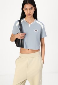 Svetlo modra kratka majica z belimi črtami na ramenih in okroglo nalepko, kombinirana s bež ožjimi hlačami in črno torbico.