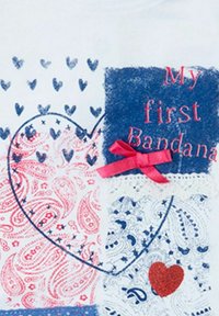 Camiseta blanca con un diseño de corazón en rojo y azul, con patrones de paisley, texto que dice "Mi primera bandana" y un acento de cinta rosa.