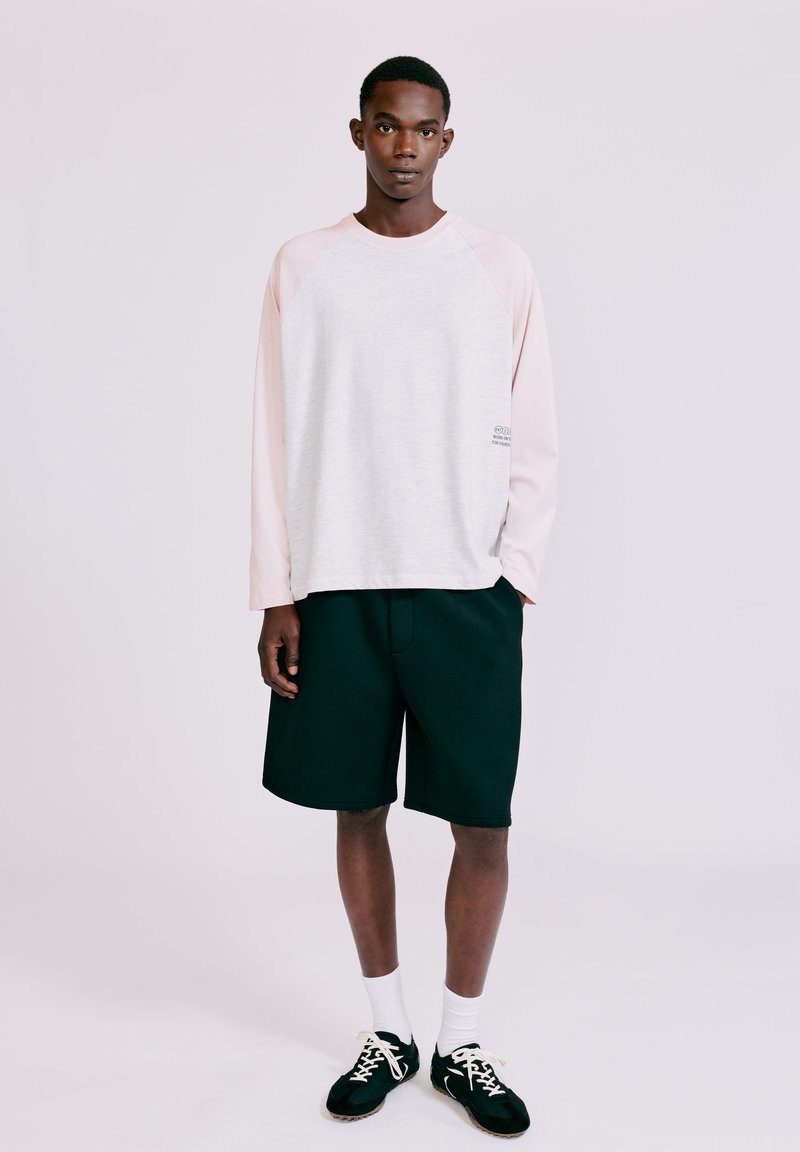 Jeune homme debout portant une chemise légère à manches longues, un short noir, des chaussettes blanches et des chaussures de sport noires sur un fond uni clair.