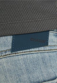 Un tejido gris texturizado se encuentra encima de una etiqueta de cuero azul con el logo en relieve "CHASIN'" sobre un denim azul claro con un sutil desgaste y costuras amarillas.