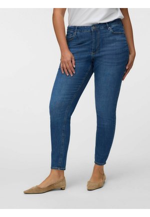 Frau trägt blaue Skinny-Jeans, weißes Oberteil und beige flache Schuhe und steht vor einem schlichten hellen Hintergrund.