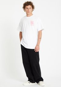 White oversized t-shirt with pink spiral graphic on the left chest, paired with loose black pants and white footwear. Smooth texture.