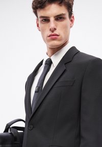 Czarny garnitur o dopasowanym kroju, biała koszula i czarny krawat z logo. Mężczyzna trzyma czarną torbę o fakturowanej powierzchni.