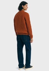 Homme avec des cheveux bouclés foncés portant un pull texturé orange brûlé, un jean bleu foncé et des baskets noires avec des semelles blanches, se tenant de côté.