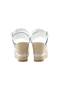 Bílé espadrilky se zkříženými popruhy, které mají tkaný juteový klínový podpatek, modro-červené pruhované akcenty a kotníkové popruhy s kovovou sponou.