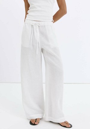 Femme portant un pantalon large en lin blanc avec une taille à cordon et des tongs noires, debout devant un fond blanc uni.