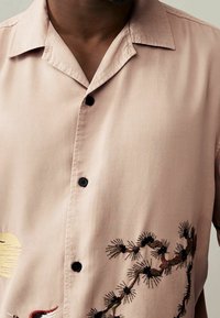 Jasnoróżowa koszula z krótkim rękawem, zapinana na guziki, z czarnymi guzikami oraz skomplikowanym haftem kwiatowym w ciemnych i jasnych kolorach.