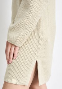 Abito in maglia beige con texture a coste, presenta uno spacco laterale e un piccolo dettaglio con etichetta all'orlo. Progettato in modo innovativo per comfort e stile.