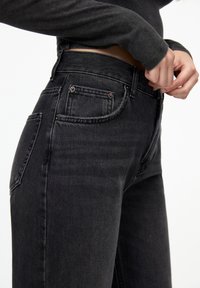 Black denim jeans with a high waist, featuring five pockets, copper hardware, and a textured fabric. Close-up view from the side.
