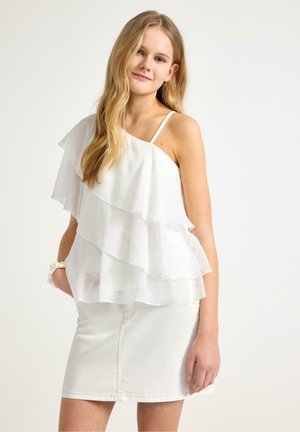 Ung kvinde med langt blondt hår iført en hvid top med flæser i lag og hvid denim nederdel, stående mod en ensfarvet baggrund.