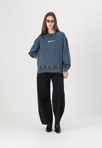 Denim sweatshirt v temno modri barvi z vstavljenimi detajli in belim logotipom. Uparjeno z črnimi širokimi hlačami in črnimi gležnjarji.