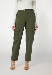 Pantaloni cargo verdi con multiple tasche frontali, una silhouette a gamba dritta e una leggera lucentezza. Abbinati a sandali dorati con cinturini.