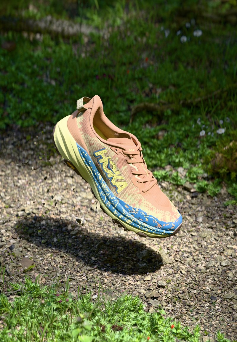 Shoe de corrida leve com uma parte superior em malha bege, entressola estampada em azul e um logótipo amarelo proeminente, projetado para uso ao ar livre.