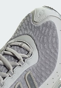 Nahaufnahme eines grauen Sneakers aus Mesh-Gewebe, mit hellgrauen Schnürsenkeln und strukturierten synthetischen Überzügen im oberen Teil des Schuhs.