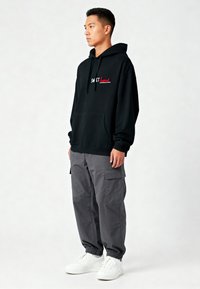 Felpa nera con cappuccio e tasca frontale, con la scritta rossa "SWEET" sul petto; abbinata a pantaloni cargo grigi larghi e sneakers bianche.