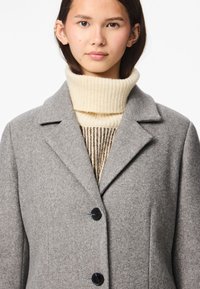 Mladá žena nosící šedý vlněný kabát přes krémový rolák, se krátkými tmavými vlasy a několika náušnicemi v uchu.