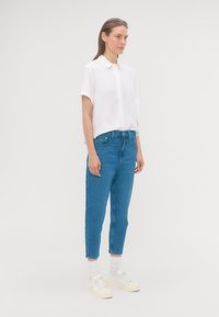 Žena stojaca v bočnom pohľade, oblečená v voľnej bielej blúzke s krátkym rukávom, modrých džínsoch s vysokým pásom, bielych ponožkách a bielych teniskách.