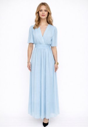 Frau steht, trägt ein hellblaues, V-Ausschnitt, kurzärmeliges, bodenlanges Kleid mit tailliertem Schnitt und schwarzen spitzen Schuhen.