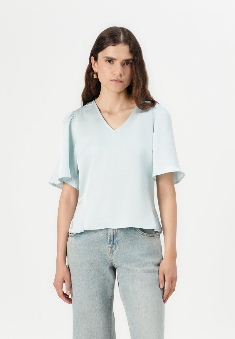 Top azzurro chiaro a maniche corte con scollatura a V e dettaglio di spalline arricciate, abbinato a jeans in denim chiaro a vita alta. Texture morbida.