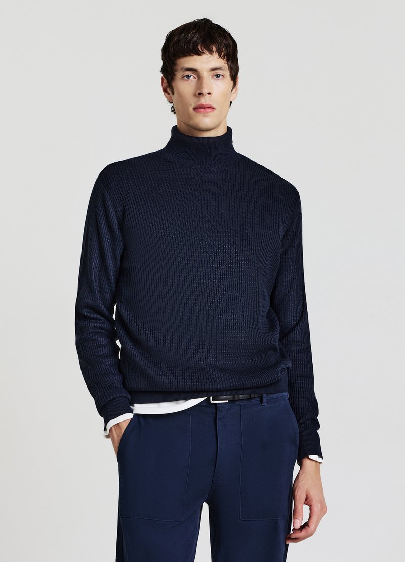 Giovane uomo che indossa un maglione a collo alto strutturato blu scuro e pantaloni blu abbinati, in piedi con una mano in tasca davanti a uno sfondo semplice.