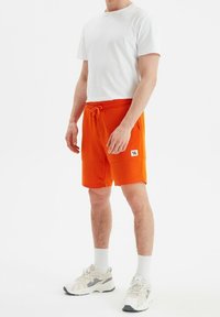 Shorts de sport orange avec une ceinture élastique et un cordon de serrage. Associés à un t-shirt blanc uni et des baskets beiges avec des accents gris.