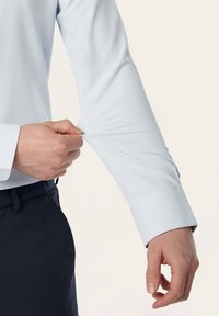 Lichtblauwe shirt met lange mouwen, gemaakt van een gladde stof en met een aansluitende manchette. De hand grijpt de mouw vast, waardoor de rekbaarheid en het ontwerp van de stof worden benadrukt.