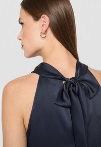 Námornícky top na zaväzovanie za krkom z textúrovanej látky s mašľou vzadu. Zlaté kruhové náušnice dopĺňajú vzhľad. Mäkk�ý lesk a elegantný dizajn.