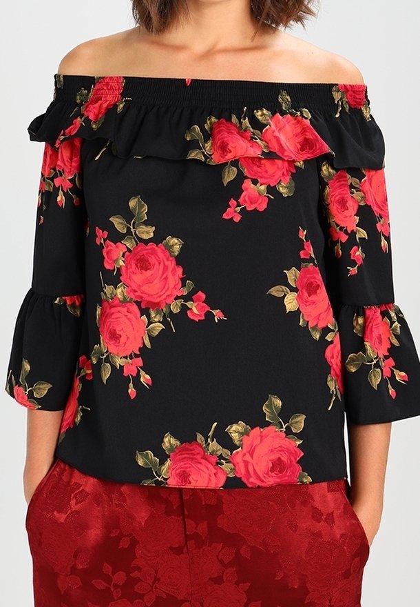 Woman wearing an off-shoulder black blouse with red rose patterns and ruffled neckline, paired with red floral-patterned pants.