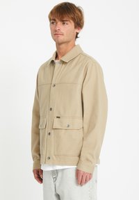 Chaqueta beige de botonadura en un corte relajado con cuello de pico, dos bolsillos frontales y una sutil textura en la tela.