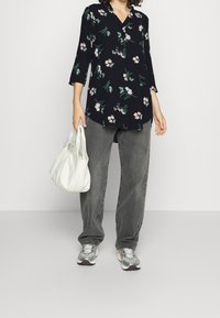 Blouse noire à fleurs avec des manches trois-quarts, associée à un jean gris ample et des baskets argentées. Tient un sac à main blanc.
