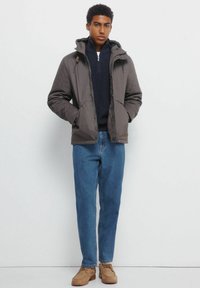 Szara, ocieplana kurtka z kapturem, zapinana na zamek z przodu, z dwoma bocznymi kieszeniami. Noszona na granatowym swetrze i zestawiona z niebieskimi dżinsami oraz beżowymi butami.