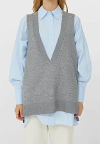 Femme portant une chemise bleu clair à boutons avec un gilet tricoté gris surdimensionné et un pantalon écru sur fond uni.