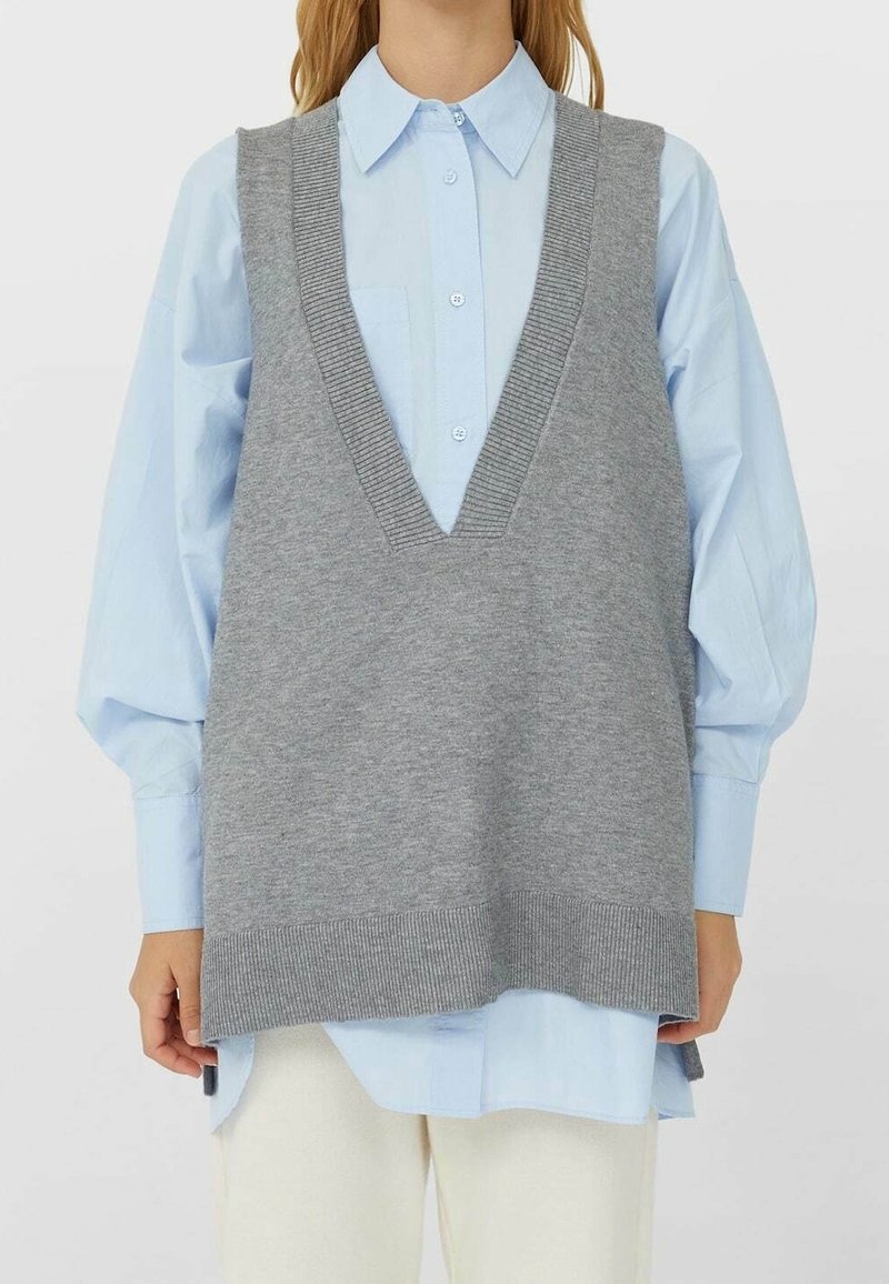 Femme portant une chemise bleu clair à boutons avec un gilet tricoté gris surdimensionné et un pantalon écru sur fond uni.