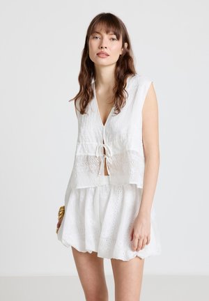 Femme aux longs cheveux bruns portant un haut blanc sans manches en broderie anglaise noué devant et une jupe blanche assortie, debout devant un fond uni.