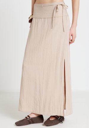 Gonna lunga beige con spacco laterale e vita annodata, indossata con ballerine marroni intrecciate, braccialetti e anelli su una persona in piedi.