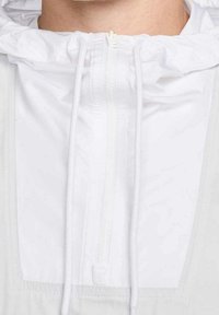 Biała bluza z kapturem, z suwakiem przy szyi, sznurkiem do regulacji kaptura oraz gładką fakturą materiału. Posiada częściowy zamek błyskawiczny z przodu oraz warstwowy design.