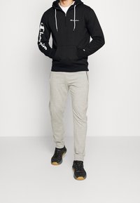 Sweat à capuche zippé noir avec logo blanc sur la poitrine et la manche, associé à un pantalon de survêtement gris clair et des chaussures noires avec une semelle en caoutchouc.