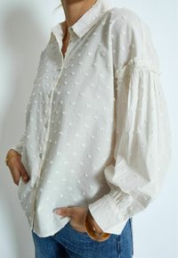 Witte blouse met knoopsluiting en textuur stippen, met geplooide mouwen en een kraag. Zachte stof met een iets ontspannen pasvorm. Gekocht in combinatie met blauwe jeans.