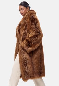 Brun faux fur frakke med løs pasform, lange ærmer og lommer. Teksturen er blød med en skinnende finish, stylet over lyse bukser.