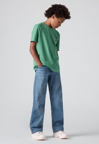 T-shirt verde de algodão com mangas curtas e detalhe de logotipo, combinado com jeans de perna larga azul claro e sapatilhas claras. Fundo liso claro.