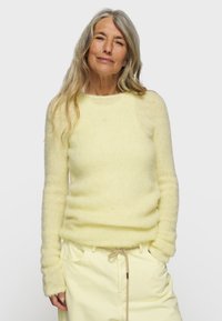 Pullover fluffy di un leggero giallo con maniche lunghe, scollatura rotonda e polsini a costine. Indossato con pantaloni leggeri gialli dotati di coulisse.