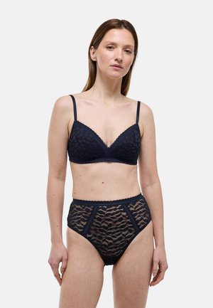 Femme portant un soutien-gorge en dentelle noire avec bretelles ajustables et une culotte taille haute assortie en dentelle, debout devant un fond clair uni.