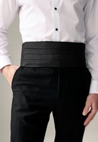 Hombre con camisa blanca de vestir metida dentro de pantalones formales negros y con una faja negra alrededor de la cintura.