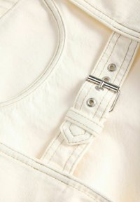 Κοντινή λήψη του λουριού από ανοιχτό μπεζ ύφασμα ενός ολόσωμου με μεταλλική αγκράφα και ενισχυμένες ραφές.