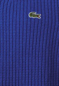 Gebreide trui in felblauw met een gestructureerd ribbelpatroon. Heeft een klein groen krokodillogo geborduurd op de voorkant.