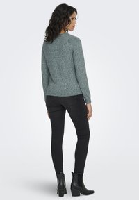 Pulover texturat gri cu guler rotund, purtat cu blugi negri, croială mulată, și cizme negre cu toc la gleznă. Văzut din spate.