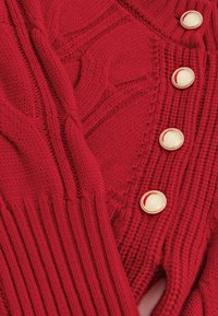 Crvena pletena kardigan jakna s teksturiranim uzorkom i rebrastim manžetama, s kremastim okruglim gumbima kao istaknutim detaljima.