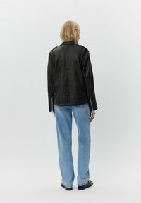 Chaqueta de cuero negra con epaulettes en los hombros, cremalleras en las mangas, acabado texturizado; combinada con unos jeans de mezclilla azul claros y zapatos negros. Vista posterior.